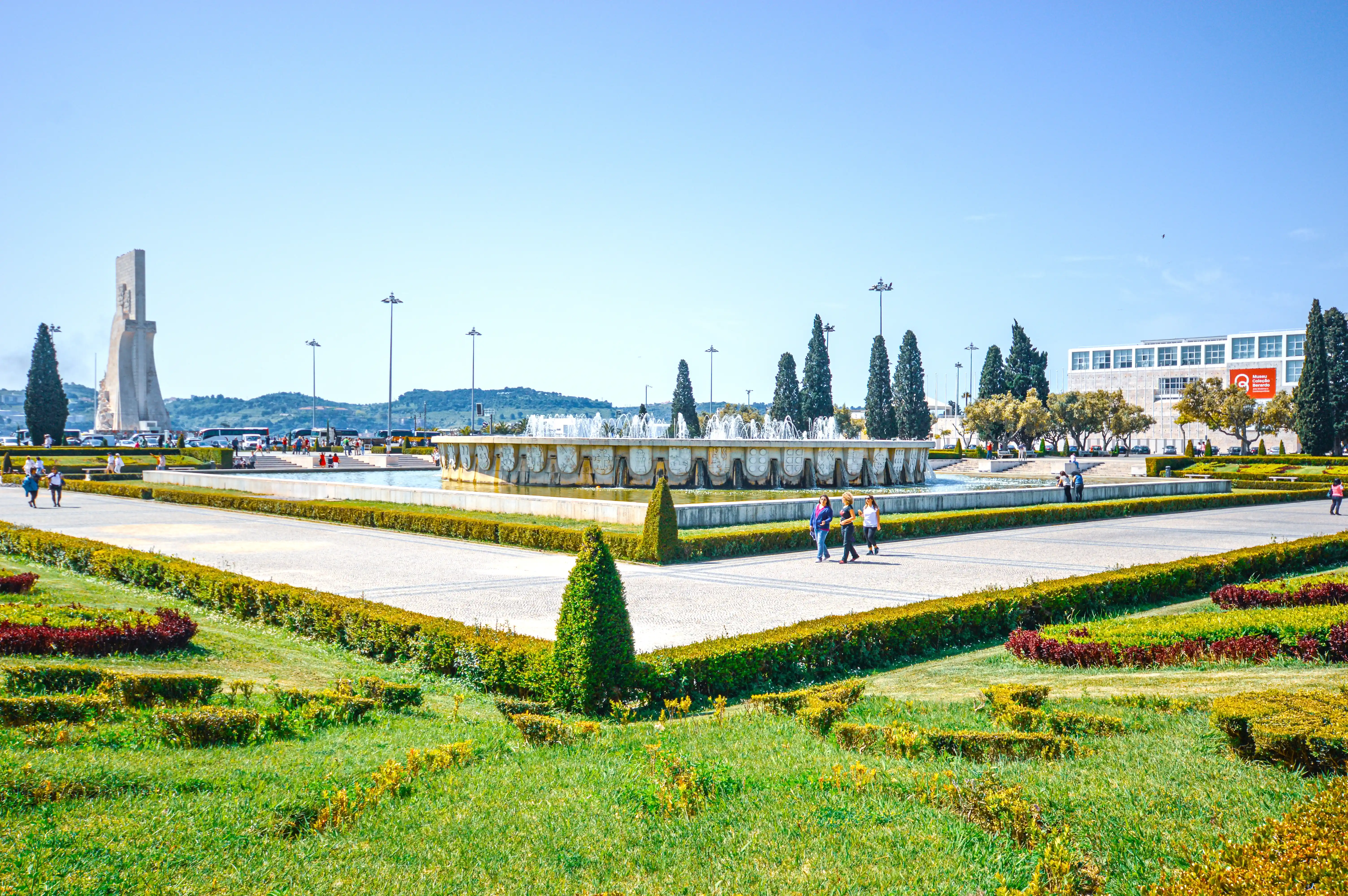 A wide view of a sunny public plaza in Lisbon, Portugal, with a large circular fountain in the centre and a tall, stone monument on the left.
